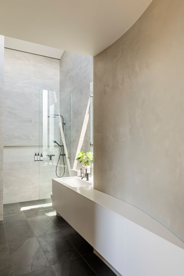 A bright, minimalist bathroom featuring a frameless glass walk-in shower, large pale stone-look wall tiles, dark grey floor tiles, and a long white floating vanity with an integrated basin. A narrow vertical window brings in natural light, while a small plant and restrained fixtures add subtle warmth.