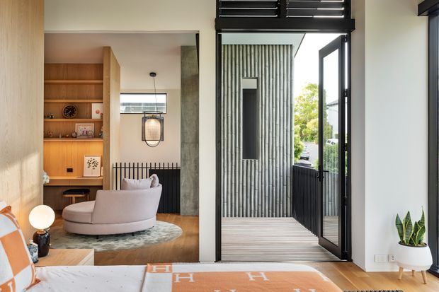 Contemporary bedroom interior opening onto a narrow timber-decked balcony or terrace. The room features warm timber built-in shelving and wall panelling, a curved upholstered lounge chair, patterned rug, pendant light, and a partially visible bed. Black-framed full-height glazing and an open glass door connect to an exterior screen-lined balcony, with a ribbed concrete or masonry wall and black louvred element above.