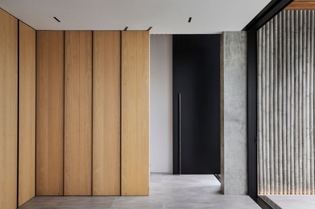 Minimalist interior entry or hallway featuring full-height vertical timber-panelled joinery on the left, a tall matte-black door, a raw concrete column, and a ribbed translucent-glass partition framed in black. The pale polished floor and white ceiling create a restrained, contemporary composition with strong linear detailing.