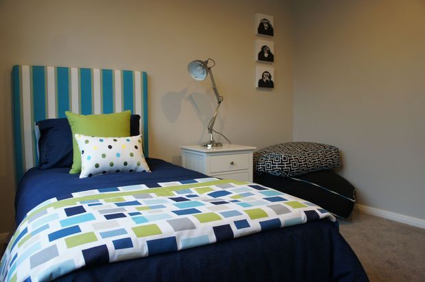 A compact bedroom with a single bed dressed in navy bedding and a bright geometric quilt featuring blue, green, white, and grey blocks. The bed has a striped blue-and-white upholstered headboard, colourful patterned cushions, and a white bedside table with a metallic adjustable lamp. A dark patterned ottoman sits beside the bed, with three small monochrome portrait artworks arranged vertically on the neutral wall.