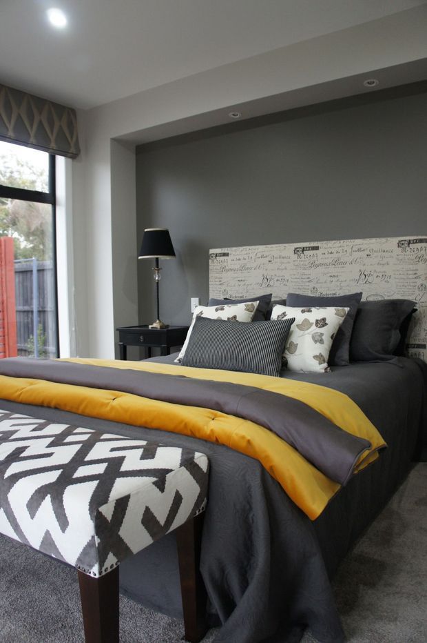 A contemporary bedroom with a dark charcoal feature wall and recessed ceiling detail behind the bed. The bed is dressed in charcoal bedding with a mustard-yellow and grey layered throw, patterned decorative pillows, and a graphic black-and-white upholstered bench at the foot. A black bedside table and floor lamp sit beside the bed, with a window and patterned Roman blind on the left.