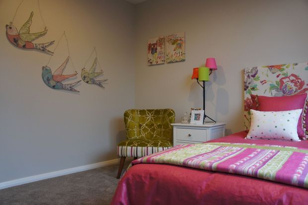 A colourful, playful bedroom with a bed dressed in bright pink, green and patterned textiles, a floral upholstered headboard, a patterned accent chair, a small bedside table and a multi-coloured floor lamp. Light grey walls feature whimsical bird artwork and children's-style paintings, creating a cheerful eclectic atmosphere.