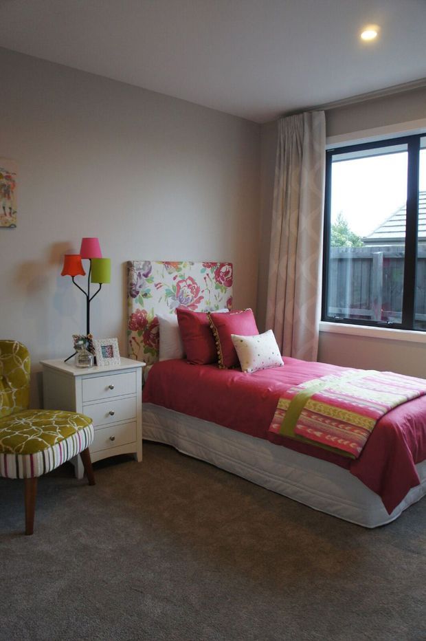 A colourful, traditionally styled bedroom with a single bed dressed in bright pink and patterned textiles, a floral upholstered headboard, a white three-drawer bedside table, and a patterned accent chair. A multi-coloured floor lamp, framed artwork, curtains, carpeted flooring, and a large dark-framed window complete the room.