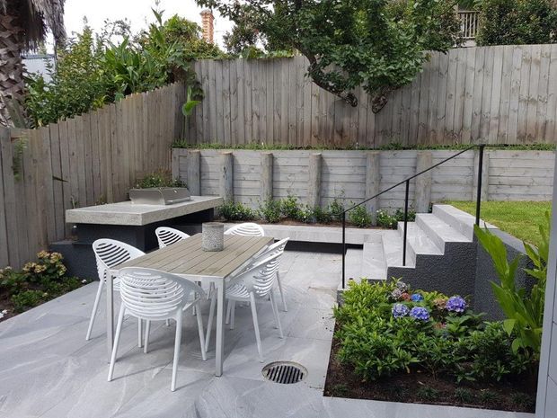 A compact landscaped backyard courtyard with a paved outdoor dining area, white outdoor table and chairs, a built-in barbecue counter, raised garden beds, timber fencing, and concrete steps with a black handrail. The space has a clean contemporary garden design with hydrangeas and other established planting.