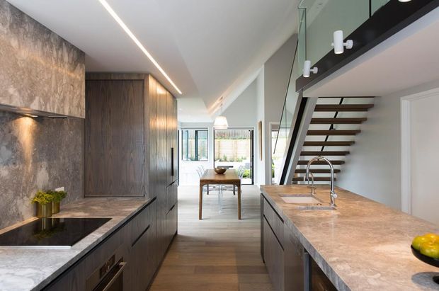 Contemporary open-plan kitchen with dark timber cabinetry, stone benchtops, integrated appliances, and a sculptural island with double sink. A dining area and bright living space extend beyond the kitchen, while an open-riser stair with a black structural stringer leads to an upper level. Linear recessed lighting and floor-to-ceiling glazing create a refined, airy interior.
