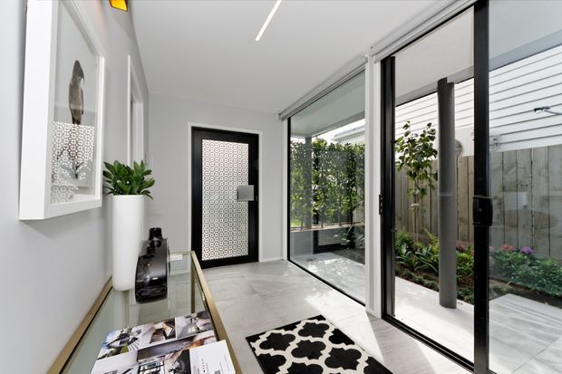 Bright contemporary entry hall with large black-framed glazed doors opening to a landscaped courtyard. The space features pale tiled flooring, white walls, a slim console, framed wall art, indoor plants, and a patterned runner, with a black-and-white patterned security or feature door at the end.