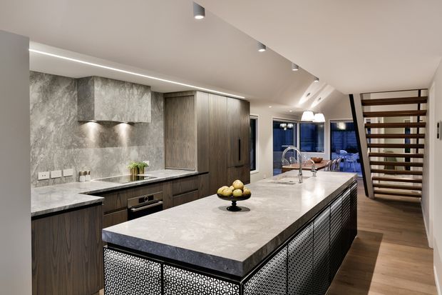 A contemporary open-plan kitchen dominated by a large stone island with integrated sink and brushed metal mesh-panelled base. Dark timber-grain cabinetry runs along the back wall beneath a grey stone splashback and rangehood, while warm timber flooring extends toward a dining or living area visible in the distance. Recessed and linear ceiling lighting create a refined, bright, high-end atmosphere. A timber slatted stair or feature screen is visible on the right.