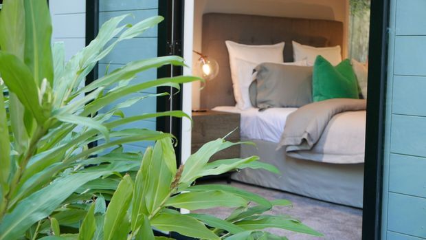 View through large glazed or open doors into a compact, warmly styled bedroom. The room features a bed with layered white and grey bedding, a padded taupe upholstered headboard, grey and green accent cushions, bedside lighting, and soft grey carpet. Large tropical foliage dominates the foreground, while blue-grey horizontal exterior cladding frames the opening.