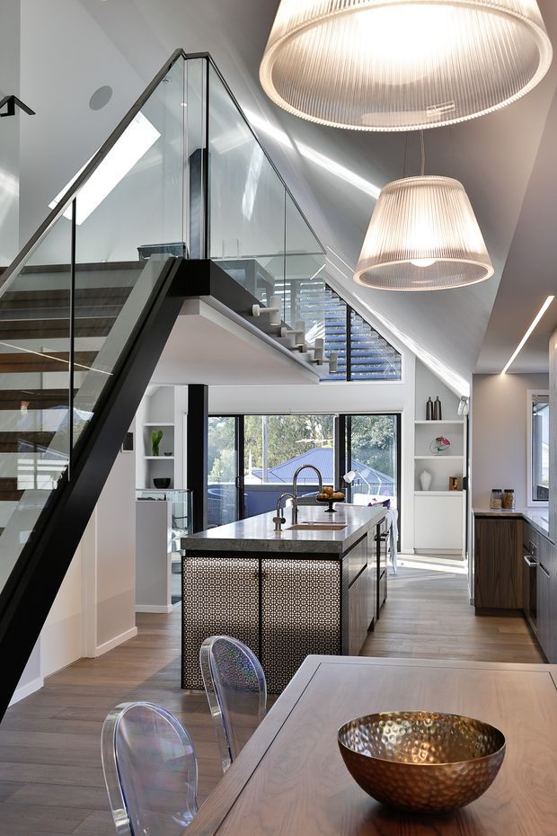 A bright, open-plan contemporary kitchen and dining area beneath a dramatic pitched ceiling. The space features a long central island with a dark stone benchtop, integrated sink and tapware, patterned decorative island panels, dark timber cabinetry, light timber-look flooring, white walls, extensive glazing, and a striking black-framed staircase with glass balustrades. Large ribbed pendant lights hang above the kitchen, while transparent dining chairs and a timber dining table occupy the foreground.