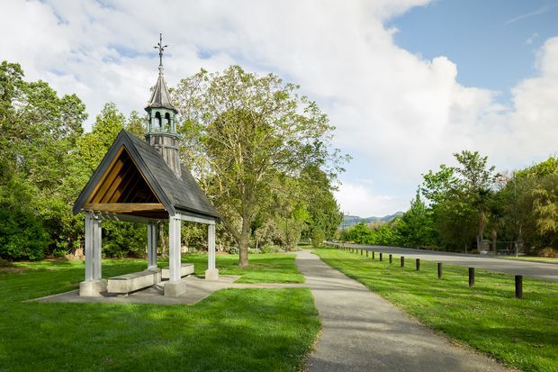 A landscaped outdoor setting featuring a small ornate timber-and-stone pavilion or chapel-like shelter with a steep gabled roof, open sides, a decorative cupola and cross finial. It sits beside a paved pedestrian path and a quiet roadway, surrounded by mature trees and manicured grass, with distant hills visible under a partly cloudy sky.