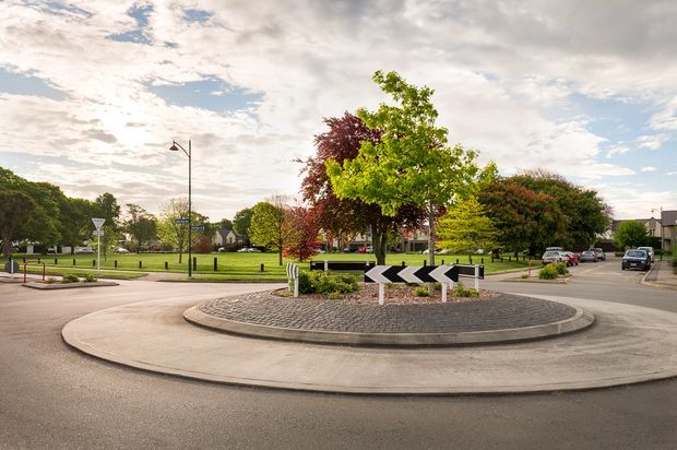 A landscaped urban roundabout with a paved circular traffic island, black-and-white directional chevrons, clipped shrubs, mature trees, street lighting, surrounding roads, and low-rise buildings under a partly cloudy sky.