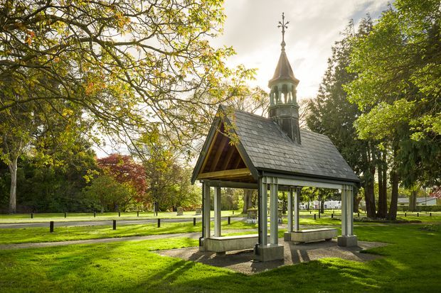 A small ornate open-sided pavilion or memorial shelter set within a landscaped park. It has a steep grey slate gabled roof, timber framing, stone or concrete posts and bases, and a decorative turret with a finial. Mature trees, lawns, paths, and park fencing surround the structure in warm late-day light.