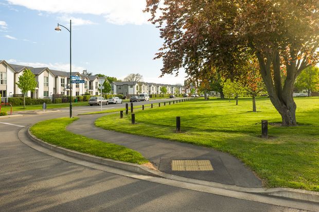 A suburban residential development viewed from a paved street, with rows of modern multi-unit homes, landscaped open green space, mature trees, pedestrian paths, bollards, street lighting, and curbs under clear daylight.