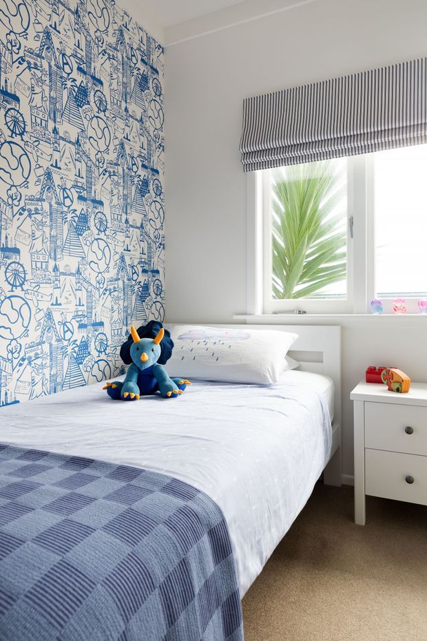 Bright children's bedroom with a single bed dressed in white bedding and a blue checked throw, a blue plush toy, whimsical blue-and-white illustrated feature wallpaper, white built-in window seating/storage, striped Roman blind, and a white bedside table. Natural light enters through the window, creating a clean, playful interior.