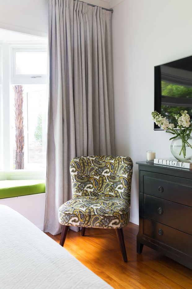 A bright, refined bedroom corner with a patterned upholstered accent chair, tall full-length grey curtains, a dark wood chest of drawers, wall-mounted television, and a window seat with a vivid green cushion. The room has white walls and warm timber flooring, creating a classic yet playful interior.