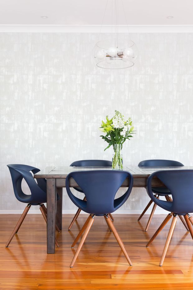 A bright dining room featuring a dark timber dining table with sculptural navy-blue chairs, polished timber flooring, a textured white wall finish, and a clear glass pendant light. A simple floral arrangement provides a small natural accent on the table.