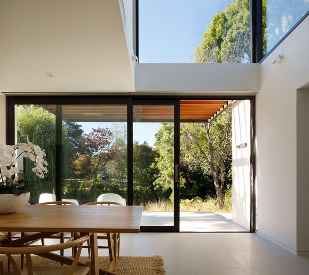 Bright contemporary dining area opening onto a landscaped outdoor terrace through large black-framed sliding glass doors. The room has a light tiled floor, white walls, a timber dining table with wooden chairs, high-level glazing, and a timber-lined covered terrace visible beyond. Abundant natural light and garden views create an airy indoor-outdoor connection.