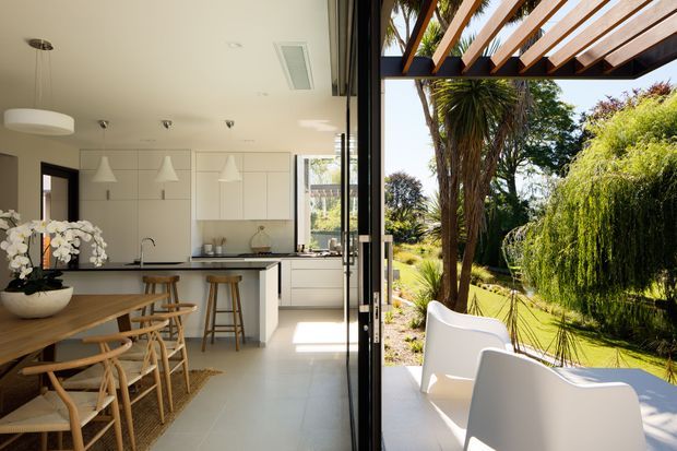 Bright contemporary open-plan kitchen and dining area opening directly onto a covered outdoor terrace and landscaped garden. The kitchen features pale built-in cabinetry, a dark stone or composite benchtop, a central island, pendant lights, and a long timber dining table with woven timber chairs. Full-height glazed doors create a strong indoor-outdoor connection. The terrace has a timber pergola with exposed beams and white outdoor seating, overlooking mature subtropical planting.