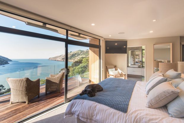 Bright contemporary bedroom with a large bed, neutral bedding, woven armchairs, timber flooring, and expansive sliding glass doors opening to a timber balcony. The room overlooks a vivid coastal bay and surrounding hills, with built-in cabinetry, a wall-mounted television, and an ensuite or dressing area visible beyond the bedroom.