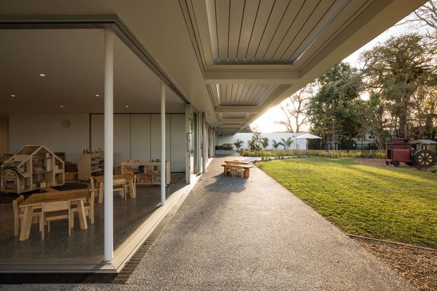 A covered outdoor walkway and terrace runs alongside a glazed, open-plan interior, with a children's play and dining area visible through floor-to-ceiling glass. The terrace has a textured paved surface, timber picnic-style tables, a slatted ceiling with recessed skylight-like openings, and slender white columns. Beyond it is a landscaped lawn and garden, creating a bright, informal connection between the building and outdoors.