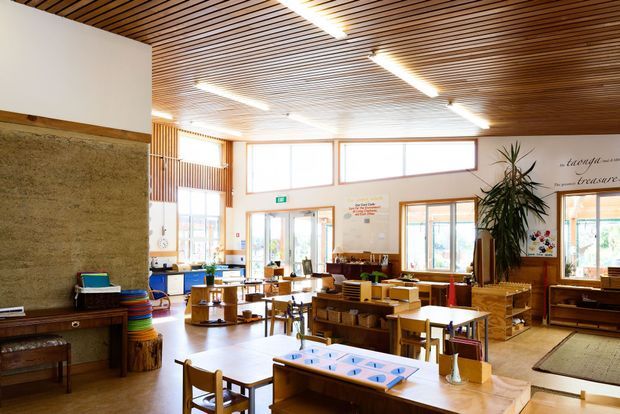 A bright, open communal interior with extensive timber slatted ceiling, large high windows, timber-framed glazing, and warm timber furniture and shelving. The space contains multiple tables and chairs, low display or storage units, a reception or service counter, indoor plants, and educational or activity materials, suggesting a childcare, community, or learning environment. A textured stone or concrete feature wall and vertical timber screening add architectural character.