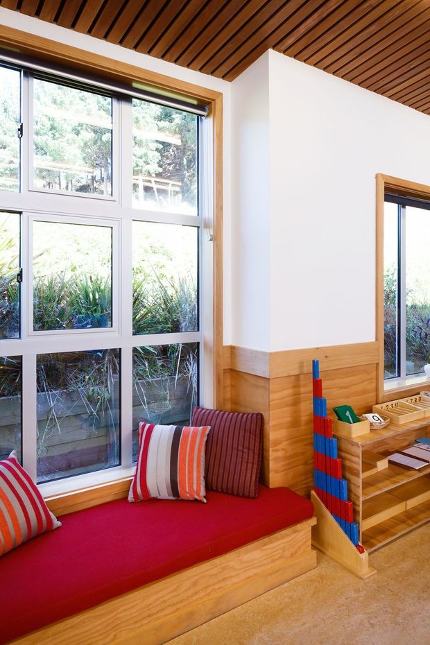 Bright, compact interior corner with large divided windows, a built-in timber window seat upholstered in red, striped cushions, and extensive custom timber joinery. The room features white walls, warm wood panelling and a slatted timber ceiling, with a built-in shelving or display unit beside the seating. Outdoor vegetation is visible through the glazing.