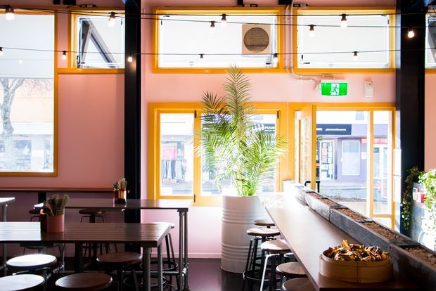 Bright industrial-style commercial interior, likely a café or casual restaurant, with a long communal counter and bar stools, dining tables, exposed ceiling services, black structural columns, and large orange-trimmed windows and doors. Pink walls, strong natural light, potted palms, and hanging string lights create a lively, warm atmosphere.