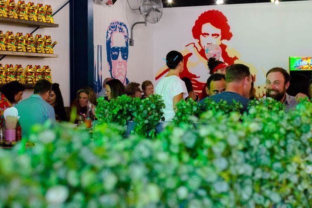 A lively commercial dining venue viewed from behind a dense green planting screen. Diners sit and stand around tables, with shelves of packaged products on the left, bright graphic wall murals, exposed services, a wall-mounted fan, and a small illuminated sign. The atmosphere is colourful, energetic, and informal.