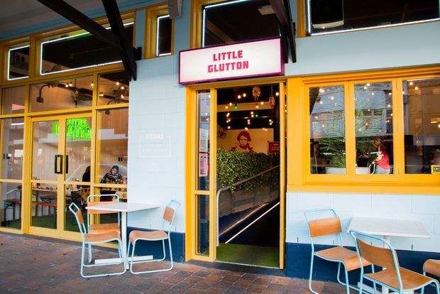 Street-facing facade of a small contemporary hospitality venue named Little Glutton, with bright yellow-orange framed glazing and doors, pale blue wall panels, outdoor café tables and mixed-color metal chairs. The open entrance reveals a warmly lit interior with plants, artwork, and pendant lights.