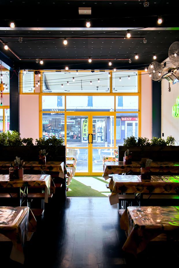 Interior view of a dark, atmospheric restaurant or casual dining venue looking toward a street-facing glazed entrance. The space has rows of timber dining tables covered with patterned tablecloths, bench seating with planter boxes, black ceilings and walls, exposed hanging pendant bulbs, wall-mounted fans, and bright yellow-orange window and door frames. A green illuminated sign is visible on the right, while storefronts can be seen through the glazing.
