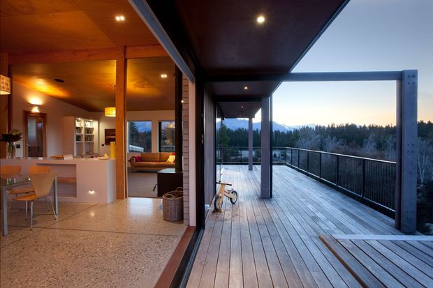 A contemporary indoor-outdoor living space photographed from a covered timber deck. The open glazing connects the deck to a warmly lit kitchen and living area, with terrazzo flooring indoors, timber ceiling lining, exposed structural posts, and black-framed glass balustrades. The deck overlooks a wooded landscape and distant hills.