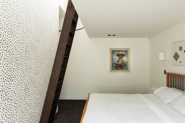 A compact contemporary bedroom with a double bed, white patterned bedding, dark carpet, and a distinctive dark timber open shelving divider or ladder-like partition. The room has off-white walls, recessed ceiling lights, framed artwork, a timber-and-black vertical slat headboard, and a wall-mounted sconce.