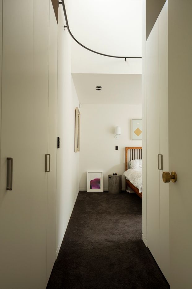 A narrow bedroom entry viewed through open double doors, with full-height white built-in wardrobes lining both sides of the passage. The room beyond contains a wooden bed with white bedding, dark carpet, a small bedside table, wall artwork, and a simple wall light. A curved black handrail or railing is visible along the upper edge, adding a distinctive architectural detail.