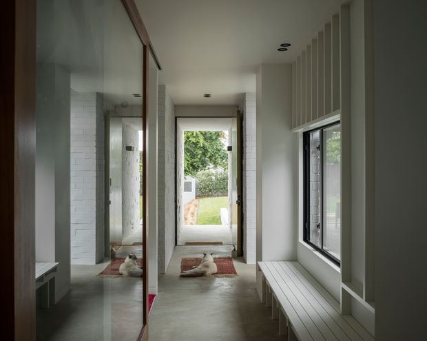 A narrow, contemporary hallway or entry corridor with polished concrete flooring, white painted brick and plaster walls, full-height dark timber-framed glazing or mirrored panels, and a built-in white slatted bench beneath a large window. Recessed ceiling lights and vertical wall battens add architectural detail. A glazed door at the end opens to a small garden, while a mirrored surface reflects the corridor and a dog.
