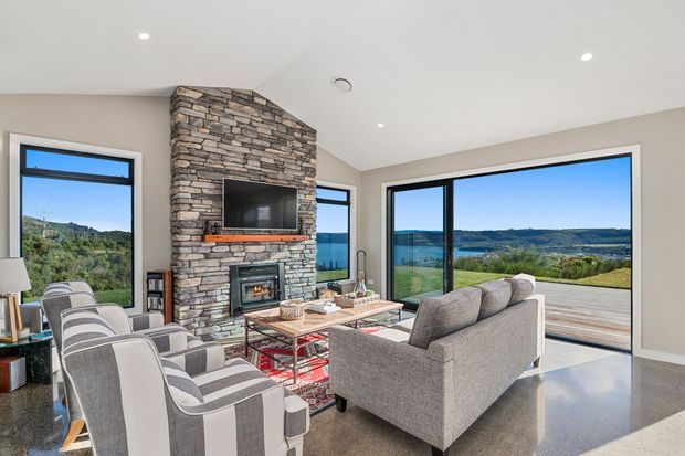 Bright contemporary living room with expansive glazing overlooking a green landscape and water. The room features a prominent stacked-stone fireplace wall with a built-in television and fireplace, a grey sofa, striped upholstered armchairs, a timber coffee table, and polished concrete flooring.
