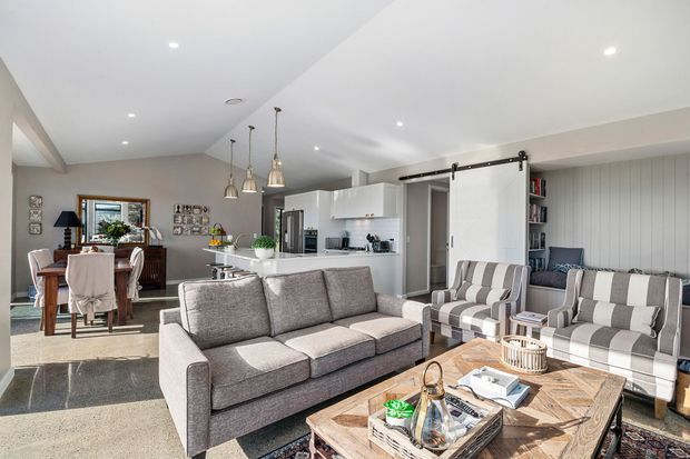 Bright open-plan living area with a large grey sectional sofa, matching striped armchairs, timber coffee table, and a white kitchen island behind it. The adjoining dining area features a timber table and chairs, while grey wall panelling, white cabinetry, pendant lights, and recessed ceiling lights create a contemporary residential interior.