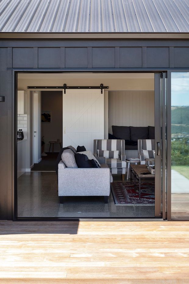 A view from a timber deck through wide sliding glass doors into a furnished living room. The room features a light grey sofa, striped armchairs, a dark sofa, patterned rug, timber coffee table, and a white barn-style sliding door. Dark painted exterior cladding surrounds the opening, with a corrugated metal roof visible above and a landscape beyond the glazing.