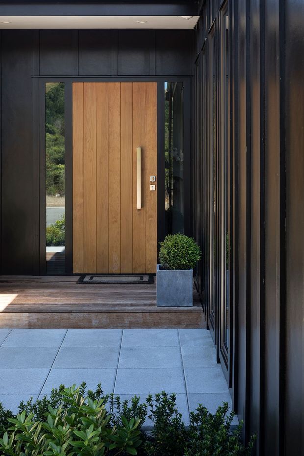 A modern residential entry viewed from the exterior, featuring a large vertical timber front door set within a dark charcoal facade. Slim dark metal framing and vertical fins line the right side, while a timber-clad porch deck, pale square paving, recessed ceiling lighting, and a square planter with a clipped shrub create a refined, sheltered approach.