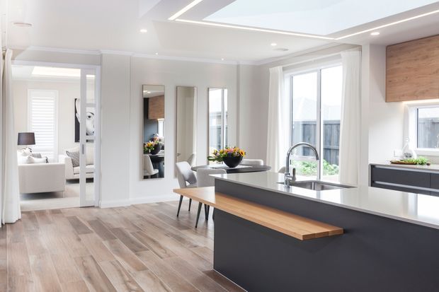 Bright open-plan contemporary kitchen with a large dark island featuring a timber breakfast ledge, integrated sink and mixer tap, pale cabinetry, and a timber-look floor. Floor-to-ceiling windows and glazed doors bring in abundant daylight, while an adjacent living area is visible through the wide opening. White walls, recessed lighting, mirrors, and restrained furnishings create a clean, spacious interior.