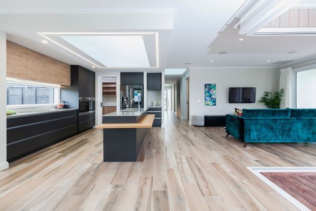Bright open-plan contemporary interior combining a modern kitchen and living room. The kitchen features dark matte cabinetry, a central island with a timber-look benchtop, integrated appliances, timber-look flooring, and a large skylight. The adjoining living area has a vivid teal sectional sofa, wall-mounted television, artwork, and indoor greenery. White ceilings, recessed lighting, and extensive glazing create an airy, spacious feel.