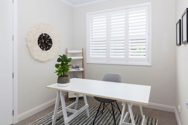 Bright, compact home office with a white trestle-style desk, grey chair, potted plant, open shelving, neutral walls, carpeted floor, and a large window fitted with white plantation shutters. Minimal wall art and clean contemporary detailing create a calm, functional workspace.