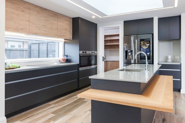 Contemporary kitchen featuring dark matte cabinetry, warm timber overhead cupboards and island seating, a pale stone benchtop, integrated appliances, timber-look flooring, and a large roof skylight. The spacious, bright room has a clean, high-contrast palette and a built-in pantry or shelving zone at the rear.
