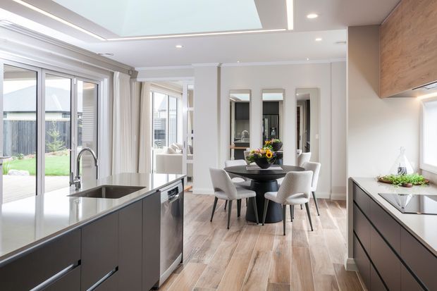 Bright contemporary open-plan kitchen and dining area with a dark island, integrated sink and appliances, light timber-look flooring, white cabinetry, and a round dining table with upholstered chairs. Large glazed doors open toward a backyard, while a skylight and recessed lighting provide abundant illumination. A timber-toned feature cabinet or wall panel adds warmth.