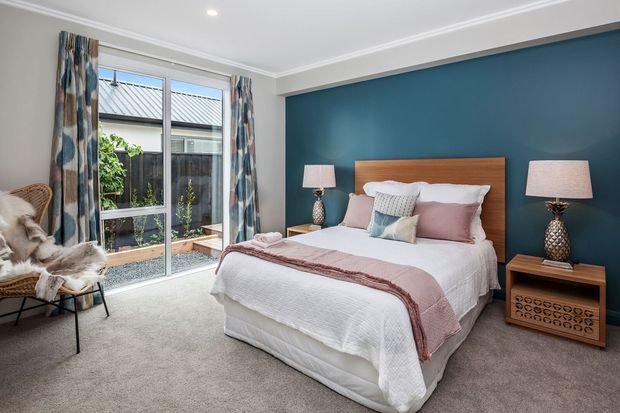 A bright, contemporary bedroom with a white upholstered bed dressed in layered white and blush bedding, positioned against a deep teal feature wall. A warm timber headboard and matching bedside tables add contrast, while patterned curtains frame full-height glazing overlooking a small courtyard. Neutral carpet, decorative lamps, and a woven lounge chair create a comfortable, refined atmosphere.