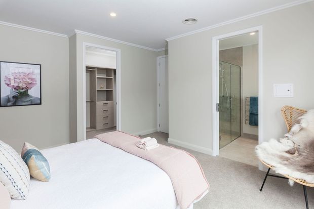 Bright, softly styled bedroom with a large bed, pale pink throw, upholstered and patterned cushions, framed floral artwork, and a woven accent chair. Built-in wardrobe storage is visible through the left doorway, while a tiled ensuite bathroom with a frameless glass shower is visible through the right doorway. The room has pale grey walls, white trim, and carpeted flooring.