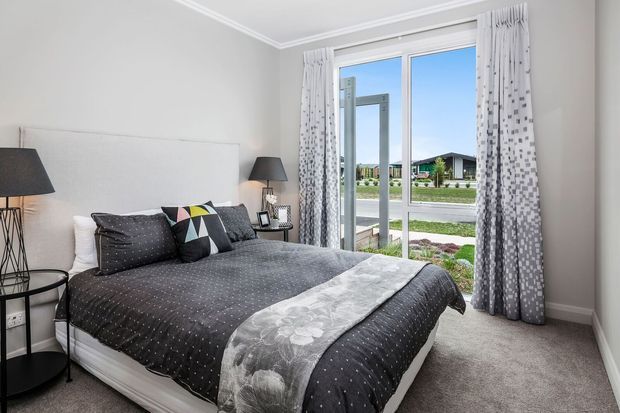 A bright contemporary bedroom with a neatly made bed, charcoal and grey patterned bedding, a geometric accent cushion, bedside tables and lamps, soft grey carpet, pale grey walls, and full-height glazed doors opening to a small outdoor area. Patterned curtains frame the view toward a suburban residential street and neighbouring homes.