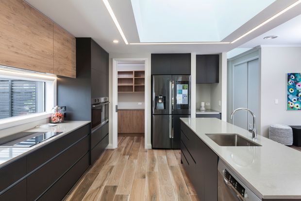 Contemporary kitchen with dark matte cabinetry, warm timber-look flooring and cabinetry, a large white island with integrated sink, black appliances, and a dramatic recessed skylight with linear LED lighting. A pantry or storage area is visible beyond the kitchen.