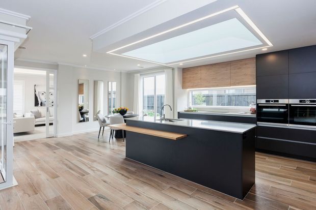 Bright contemporary open-plan kitchen with a large dark island, integrated black cabinetry and appliances, timber-look flooring, pale walls, and a striking recessed ceiling light feature. A dining area and living space are visible beyond the kitchen, with abundant glazing providing natural light.