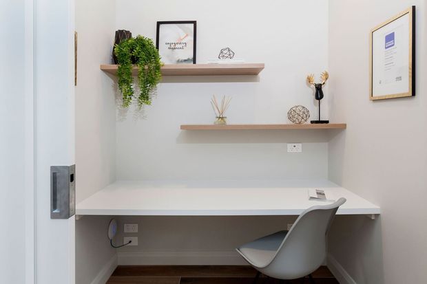 Compact built-in home workspace with a white desk spanning a recessed alcove, a modern molded chair, two floating timber shelves, framed artwork, decorative objects, and trailing greenery. The space has pale grey walls, dark timber flooring, and a clean, contemporary minimalist appearance.