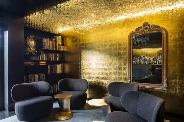 A dramatic, luxurious lounge and home bar interior with deep charcoal walls, built-in bookshelves, sculptural dark upholstered lounge chairs, and small metallic side tables. A highly reflective gold-toned textured ceiling and feature wall create a warm glow, while an ornate framed mirror reveals a compact bar with bottle shelving and stools behind it.